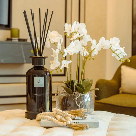 A decorative display features an Extra Large Luxury Sable Onyx Room Diffuser, a glass vase with white orchids, two stacked books, and a string of wooden beads on a tufted ottoman. The background includes a green armchair and a minimalist interior setting.