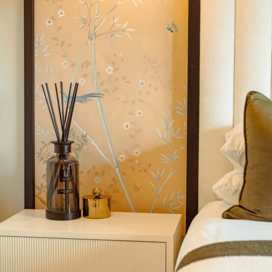 A soothing bedroom scene features a beige nightstand adorned with the Extra Large Luxury White Musk Room Diffuser in a dark glass bottle and a small golden container. Behind, a wall panel painted with delicate botanical designs enhances the tranquil atmosphere. A part of a neatly made bed with white and green pillows is visible in the background.