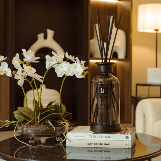 A cozy living room scene features a round glass table adorned with an arrangement of white orchids and an Extra Large Luxury White Musk Room Diffuser with reeds. The table also holds two books titled 