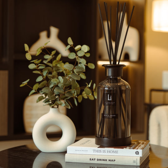 A stylish home interior features an Extra Large Luxury White Musk Room Diffuser with black reeds and a bottle labeled "Li" next to a minimalist white vase holding green foliage. Three books titled "THIS IS HOME," "FK PLASTIC," and "EAT DRINK NAP" are stacked beside the vase, adding a decorative touch.
