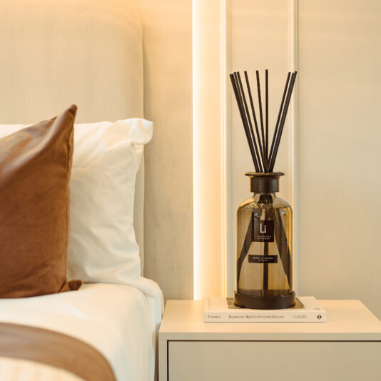 A bedside table featuring an Extra Large Luxury White Musk Room Diffuser and two stacked books on top of it. The adjacent bed is adorned with white sheets and a brown pillow. The room exudes a warm and cozy ambiance, enhanced by soft lighting.