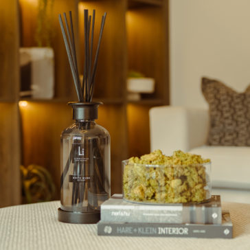 Extra Large Luxury White Musk Room Diffuser A cozy living room scene features the Extra Large Luxury White Musk Room Diffuser with dark sticks on a circular wooden tray atop a stack of three interior design books. Beside the diffuser, there is a transparent bowl filled with green dried moss. In the background is a comfortable white sofa.