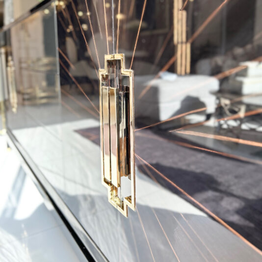 Close-up of a decorative, sunburst-like pattern centered around a sleek, golden door handle with geometric design, reflecting light on a shiny black surface in a modern interior setting. The High End Veneer Art Deco Style Sideboard adds an opulent touch to the shadowy room in the background.