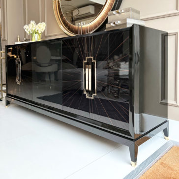 High End Veneer Art Deco Style Sideboard The High End Veneer Art Deco Style Sideboard, featuring stylish handles and elegant gold accents, stands against a wall. A round mirror and two decorative objects rest on top, while white flowers in a vase add a fresh touch. The scene is set in a pristine, modern interior space.