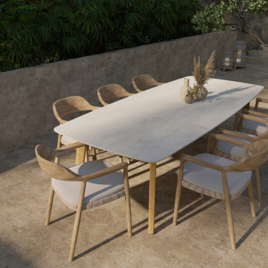 A modern outdoor dining area featuring a rectangular stone table surrounded by eight Outdoor Teak Woven Modern Dining Chairs, each equipped with light grey cushions. Neutral-colored vases and dried flowers embellish the table, while the space is enclosed by a concrete wall and lush green foliage.