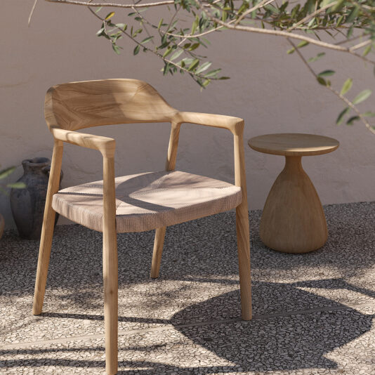 An Outdoor Teak Woven Modern Dining Chair with a minimalist design sits on a pebbled surface next to a small, rounded wooden side table. They are positioned under the shade of leafy branches against a light-colored wall, with a couple of ceramic pots visible in the background.