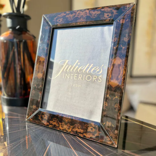 A decorative black and brown patterned picture frame, part of the Set of 3 Burr Walnut Effect Photo Frames, is placed on a reflective surface with the label 