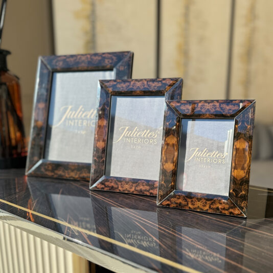 A set of 3 Burr Walnut Effect Photo Frames of varying sizes is elegantly placed on a glossy black and white surface. Each frame contains a placeholder with the text "Juliettes Interiors" and different size labels. In the background, there's a blurry brown and cream-colored wall.