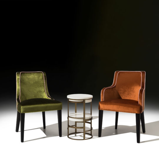 Two elegant High End Occasional Chairs With Piping Detail and velvet upholstery, one in olive green and the other in burnt orange, are placed on a white floor. A small round side table with a white tabletop and gold metal frame stands between them. The dark background creates a striking contrast.