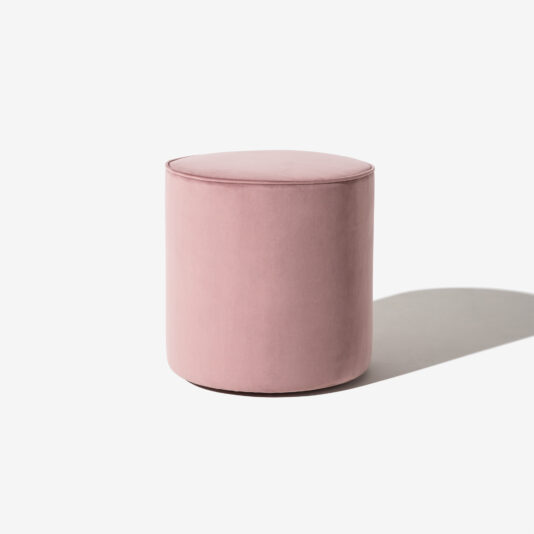 A High End Round Pouffe, upholstered in soft pink velvet, is pictured against a plain white background. The ottoman casts a shadow to its right, emphasizing its cylindrical shape.