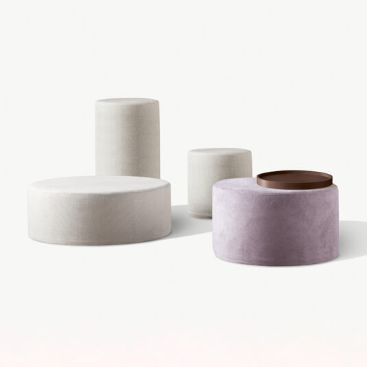 Against a white background is a minimalist display of four differently sized and colored cylindrical ottomans. The High End Round Pouffe, in light gray, stands out prominently among them. Another ottoman is light beige, while the two lavender-toned ones complete the arrangement, with one adorned by a round brown tray on top.