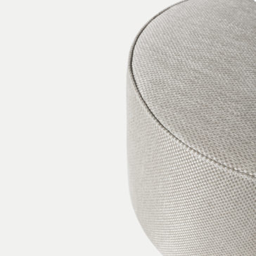 High End Round Pouffe Close-up of the High End Round Pouffe, revealing its light gray textured fabric with an intricate weave and slightly curved surface. The subtle gradient and fine detailing of the material are highlighted, emphasizing its elegant and smooth finish—ideal for this high-end piece.