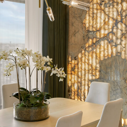 Elegant dining room in a chic Battersea apartment featuring a light wood table with white upholstered chairs. A glass vase with white orchids is centered on the table, while a marble accent wall with warm backlighting and a modern gold chandelier enhance the room's ambiance.