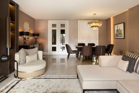 Elegant living and dining area with beige and gold decor, reminiscent of a meticulous design stage. Features a round chair, sectional sofa, glass-top dining table with six chairs, a chandelier, and a console with lamps. Large double doors and a monochrome framed photo complete the sophisticated setting.