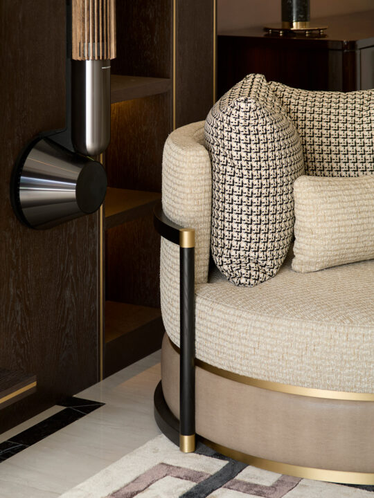 Luxurious round chair with beige and black houndstooth cushions, featuring gold and black metal accents, exemplifies the design stage mastery. It's placed near a wooden bookshelf and a modern wall light, resting on a patterned rug.