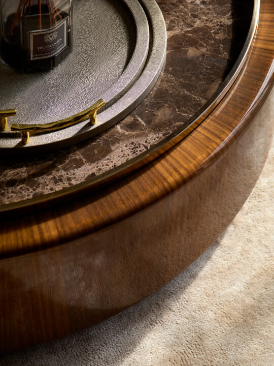 A close-up of a round, polished wooden table, still in the design stage, features a circular stone inlay with a gold handle. Atop it sits a modern bottle of fragrance. The floor beneath showcases a textured beige rug.