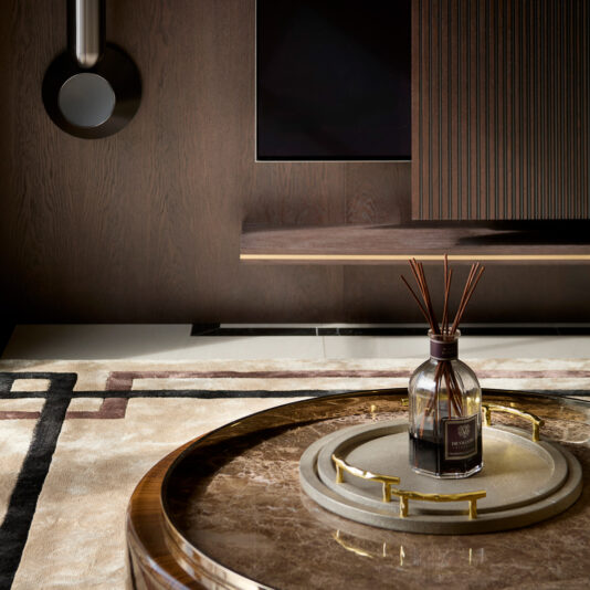 A modern luxury living room setting captures the design stage with a circular wood and marble coffee table. An aroma diffuser with wooden reeds sits on the table, set against a backdrop of wood-paneled walls and a mounted light fixture. The area rug boasts a geometric design.
