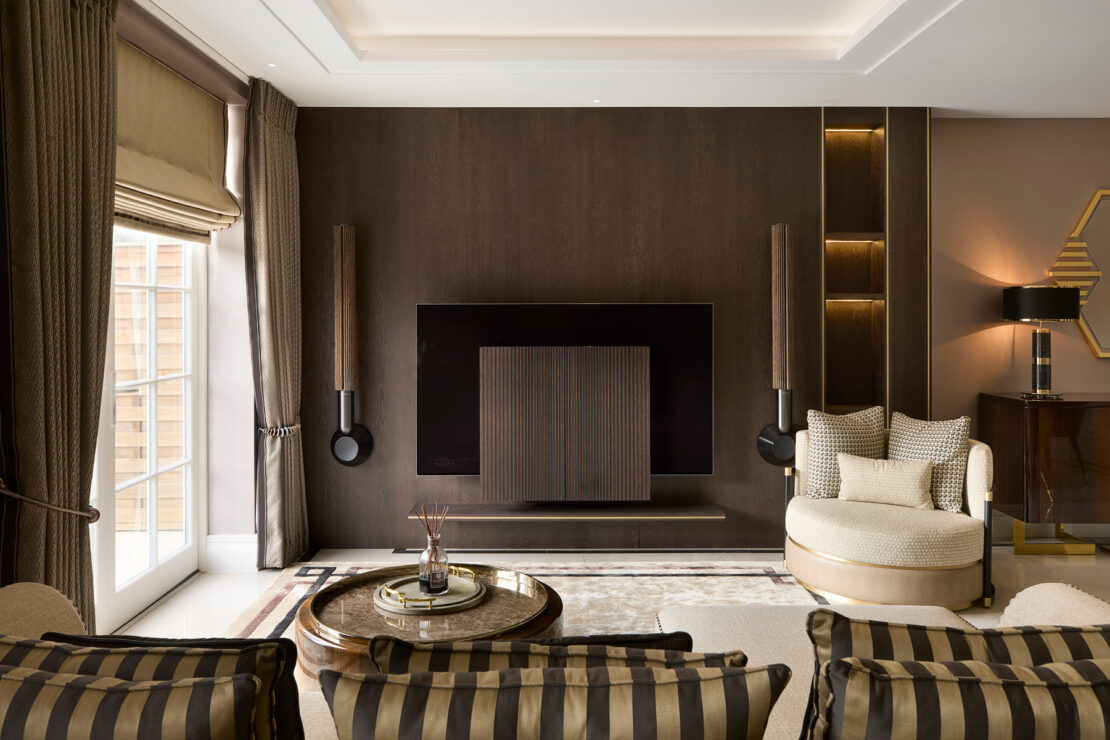 Modern living room with a large TV mounted on a dark wooden accent wall, flanked by two vertical speakers. A round armchair with plush cushions and striped chairs face the TV. Floor-to-ceiling windows fill the space with natural light, highlighting stylish decor elements reminiscent of a design stage.