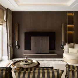 Modern luxury living room design project by Juliettes Interiors. With a large TV mounted on a dark wooden accent wall, flanked by two vertical speakers. A round armchair with plush cushions and striped chairs face the TV. Floor-to-ceiling windows fill the space with natural light, highlighting stylish decor elements reminiscent of a design stage.