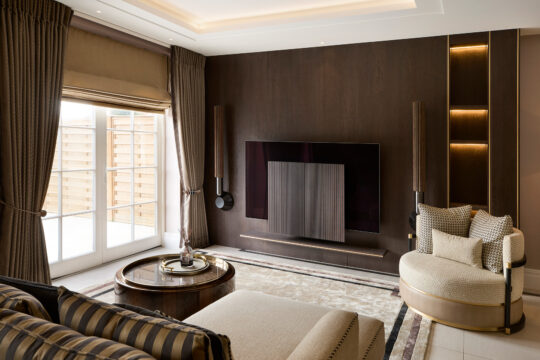 A cozy living room in the design stage boasts a modern touch with a large flat-screen TV mounted on a dark wooden panel wall. It includes a round glass coffee table, beige sofa, patterned armchair, and floor-to-ceiling windows with curtains. Soft lighting adds warmth.