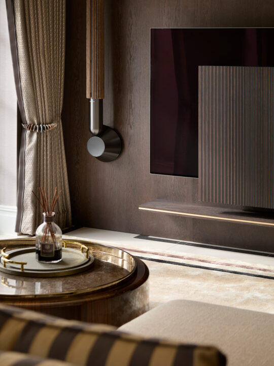 A Luxury Living Room corner with beige and gold curtains, a modern dark wood wall with a mounted flat-screen TV, a round gold coffee table with a glass bottle, and a beige armchair accented by a striped pillow.