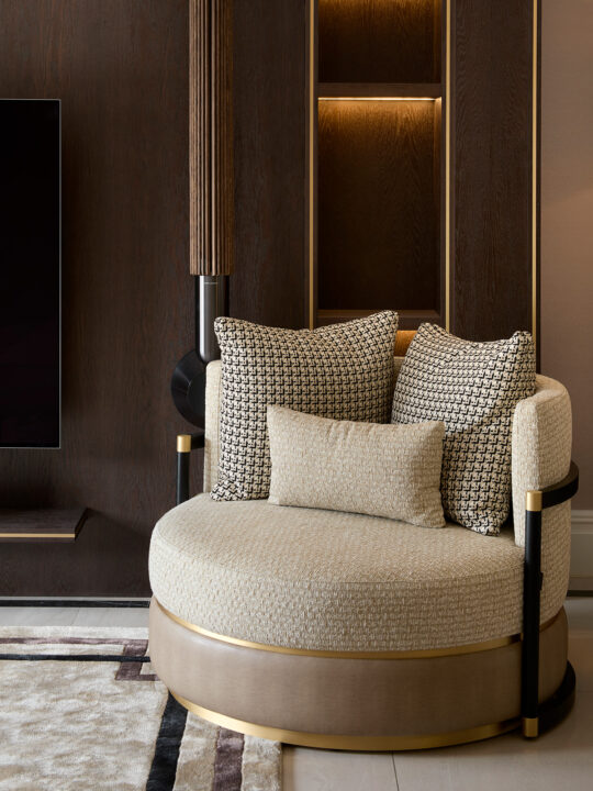 Round beige armchair with cushioned backrest and geometric-patterned pillows, placed against a dark wooden wall with built-in shelves. Subtle lighting enhances the cozy, modern design, as if perfectly set in a design stage.