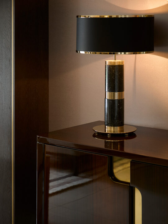 A luxurious table lamp with a black shade and a gold and black marble base sits on a polished wooden cabinet in a dimly lit room. In its final design stage, the lamp casts a warm glow on the wall and furniture, creating an elegant ambiance.