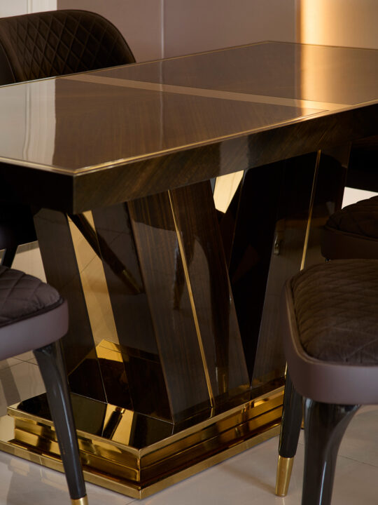 A modern dining table with a glossy dark wood surface and a geometric, gold-accented base showcases the pinnacle of design stage elegance. Four dark upholstered chairs with curved backs and quilted fabric encircle the table, exuding luxurious contemporary style.
