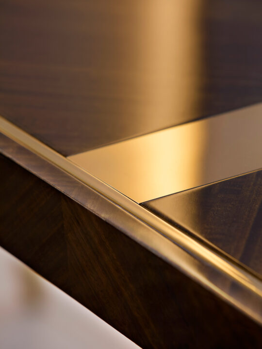 A close-up of an elegant wooden table, still in its design stage, features a polished gold inlay reflecting a warm glow. Its smooth surface and geometric pattern convey a sense of luxury and craftsmanship.