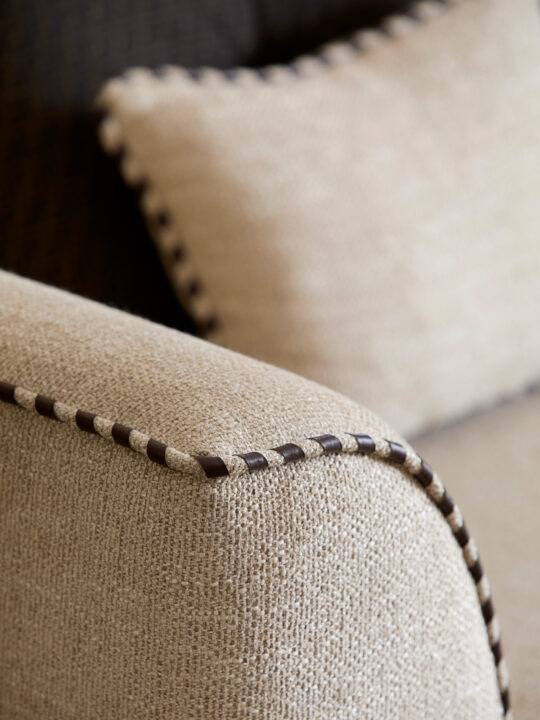 Close-up of a beige fabric armchair with a textured surface, showcasing the attention to detail often emphasized in the design stage. The armrest features dark brown stitching along the edge, while a matching cushion with similar stitching appears in the background.