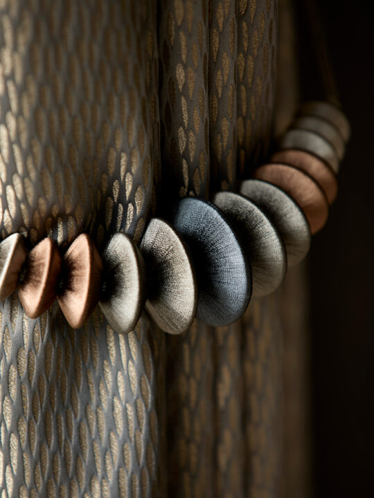 Close-up of a decorative curtain tieback with oval beads in shades of bronze, gray, and silver against a gold-patterned, dark fabric curtain. The lighting highlights the textures and colors of the beads and fabric, akin to elements carefully chosen at the design stage.