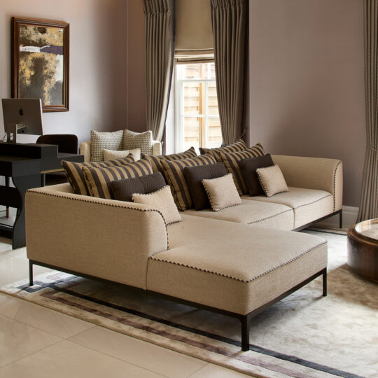 A modern living room features a beige sectional sofa adorned with striped and solid cushions. In the background, a computer rests near large artwork, suggesting a creative design stage. Floor-to-ceiling curtains and a patterned rug complete the elegant setting.