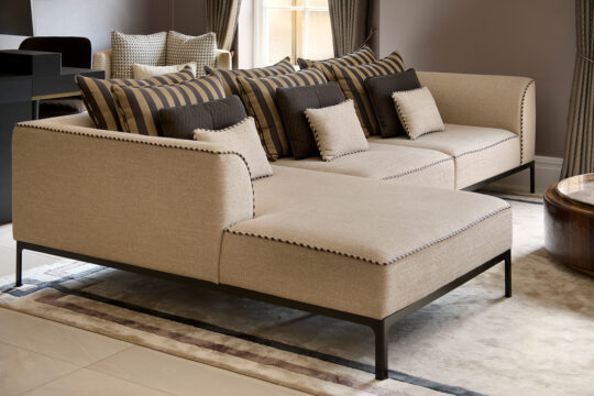 A modern beige sectional sofa with a chaise lounge takes center stage in this stylish living room, embodying the essence of refined design. It boasts striped and solid cushioned pillows, enhanced by a window with curtains and a round wooden coffee table on a patterned rug.