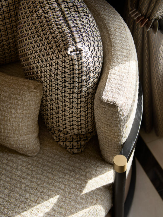 Close-up of a stylish modern armchair in sunlight, showcasing a houndstooth-patterned backrest, beige cushions, and a textured seat. A metal frame with gold accents outlines the chair, reflecting its design stage while complementing the neutral tones.