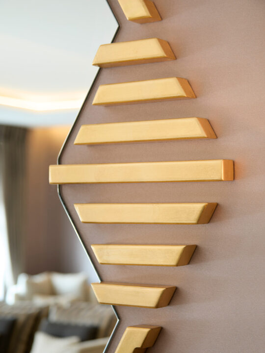 Close-up of a modern decorative wall feature with a zigzag pattern crafted from gold bars, showcasing its design stage brilliance. The background reveals a blurred interior with neutral-toned furniture and soft lighting.