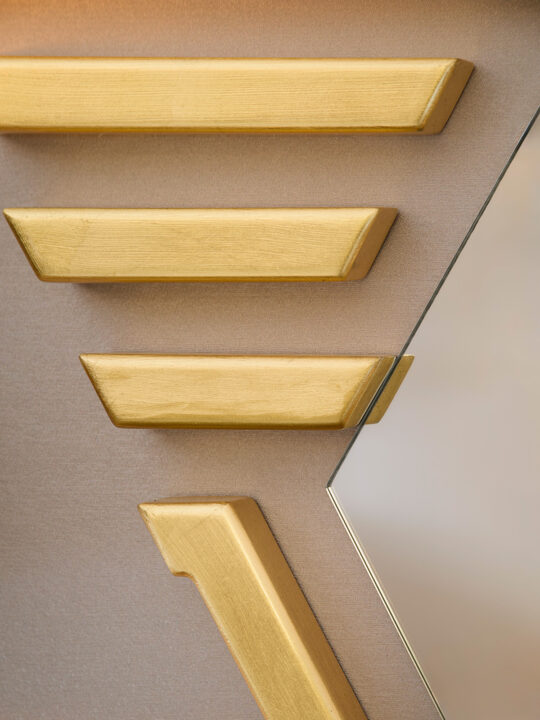 Close-up of a geometric wall design, reminiscent of a design stage, featuring gold-colored triangular and rectangular accents against a textured beige background. The composition includes a partial view of a mirror with a slanted edge.