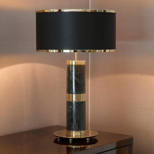 A modern table lamp with a black cylindrical base featuring gold accents and a round black lampshade stands as if in its final design stage. It gracefully rests on a polished dark wood surface against a beige wall.