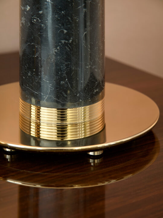 At the design stage, this close-up showcases a dark marble cylindrical object with gold banding at its base, elegantly resting on a shiny gold circular platform atop glossy dark wood.