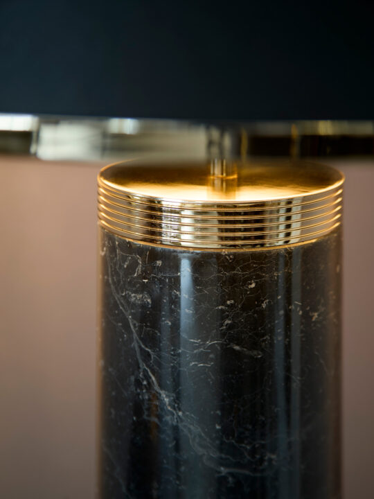 Close-up of an elegant table lamp, expertly crafted in the design stage, featuring a cylindrical black marble base with subtle veining, topped by a shiny gold accent and a dark lampshade above.