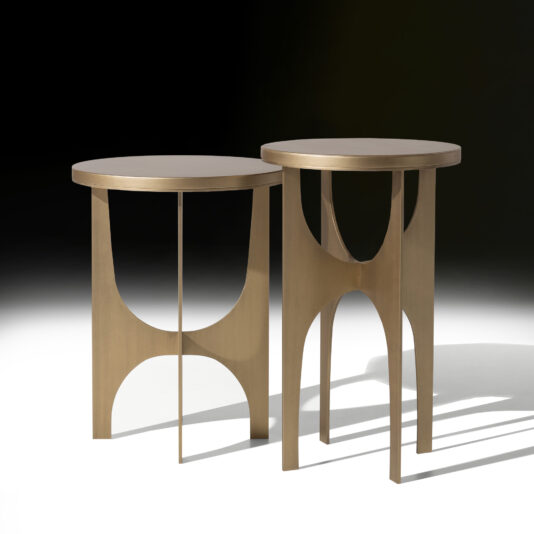 Two Art Deco Style Round Side Tables, boasting a modern minimalist design, are elegantly set against a dark background. Crafted from light wood, each table features a round top and uniquely arched legs that evoke an open, airy aesthetic. The lighting accentuates their smooth, polished surfaces and distinctive angular cuts.