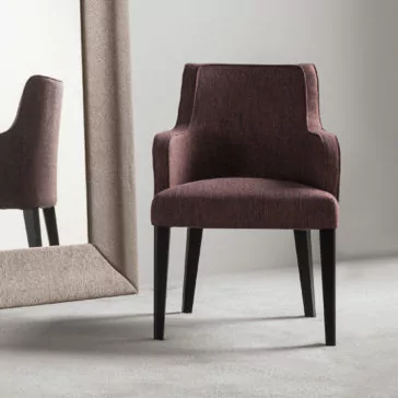 The High End Occasional Armchair, adorned in luxurious maroon fabric and supported by sleek black legs, is placed on a light gray carpeted floor. It faces a large rectangular mirror with a beige frame that partially reflects the chair. The backdrop showcases a minimalist gray wall.