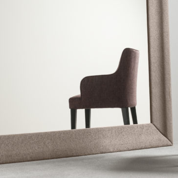 A High End Occasional Armchair with tall black legs is reflected in a large, angled mirror. The chair, set against a plain off-white background, enhances the minimalist aesthetic. The mirror features a thick beige frame.