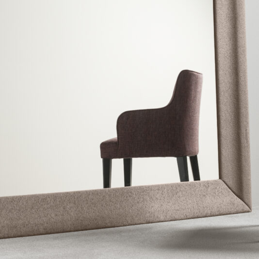 High End Occasional Armchair A High End Occasional Armchair with tall black legs is reflected in a large, angled mirror. The chair, set against a plain off-white background, enhances the minimalist aesthetic. The mirror features a thick beige frame.