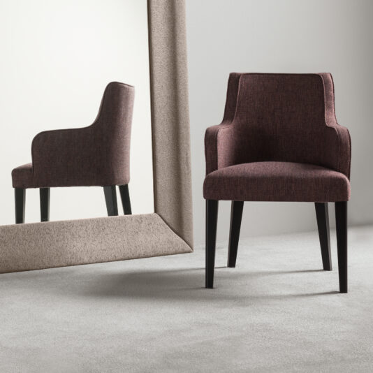 High End Occasional Armchair A High End Occasional Armchair with dark wooden legs stands in front of a large, framed mirror. The chair's modern, burgundy fabric contrasts against the light grey carpet and white wall, creating a minimalist room setting.