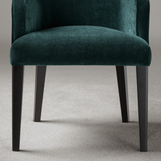 A close-up of the High-End Occasional Velvet Armchair in dark green, showcasing its luxurious design with sleek black legs, positioned on a light gray carpeted floor. The chair displays an elegant curved backrest and a plush, cushioned seat against a plain, neutral gray wall.