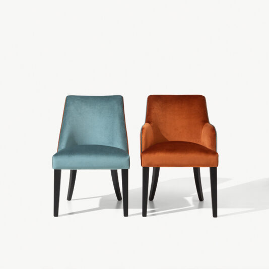 Two High End Velvet Dining Chairs With Piping sit side by side against a white background. The chair on the left is blue, while the chair on the right is orange. Both chairs have black legs and slightly curved armrests, exuding elegance and sophistication.