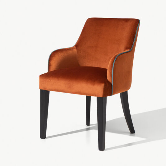 Introducing the High End Velvet Dining Chair With Piping: a modern chair adorned with burnt orange velvet upholstery and sleek piping, featuring a curved backrest and armrests. This high-end dining chair boasts black wooden legs and a sleek design, making it perfect for both dining and accent seating. The background is plain white.