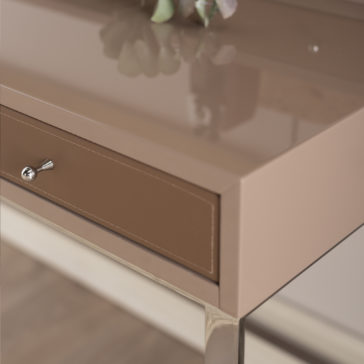 Lacquered Designer Writing Desk Close-up of the Lacquered Designer Writing Desk, showcasing its modern, sleek beige finish and single drawer with a small metallic knob. A portion of a leafy plant is slightly blurred resting on the desk's glossy surface, while the background remains out of focus to emphasize the details of the desk.