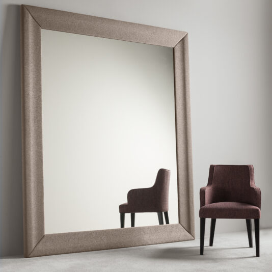 A Large Contemporary Fabric Floor Mirror with a thick, beige frame leans against a plain, light gray wall. In front of the mirror stands a small, burgundy upholstered chair with black legs, its reflection clearly visible in the mirror's surface.