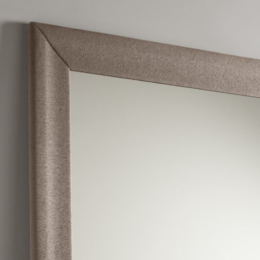 Close-up of the upper corner of the Large Contemporary Fabric Floor Mirror with a textured, metallic frame. The mirror reflects a soft, neutral off-white wall. The frame has a subtle, light beige tone with a smooth, slightly shimmery finish.
